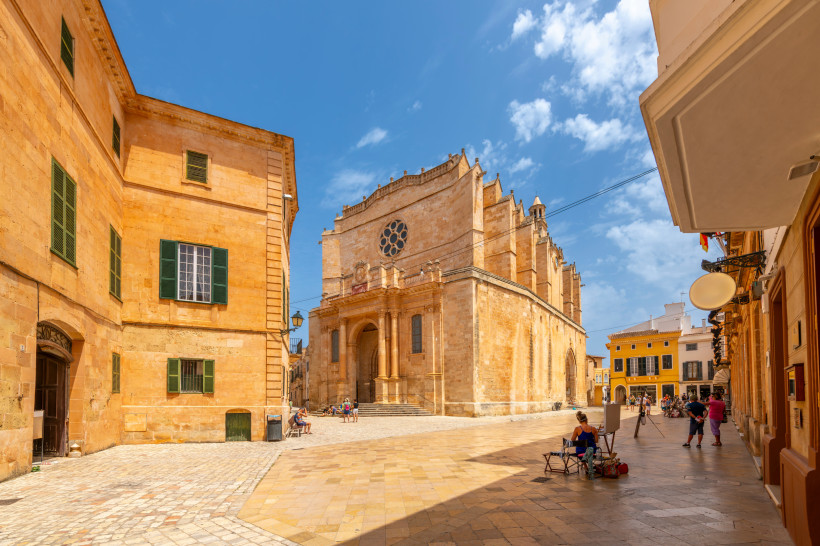 Helle Sandsteinfassaden umgeben einen sonnigen Platz in Ciutadella auf Menorca. Im Zentrum steht die beeindruckende Kathedrale Santa Maria mit gotischer Fassade und einem großen Rosettenfenster. Auf dem Platz sitzen Menschen entspannt im Schatten, einige 