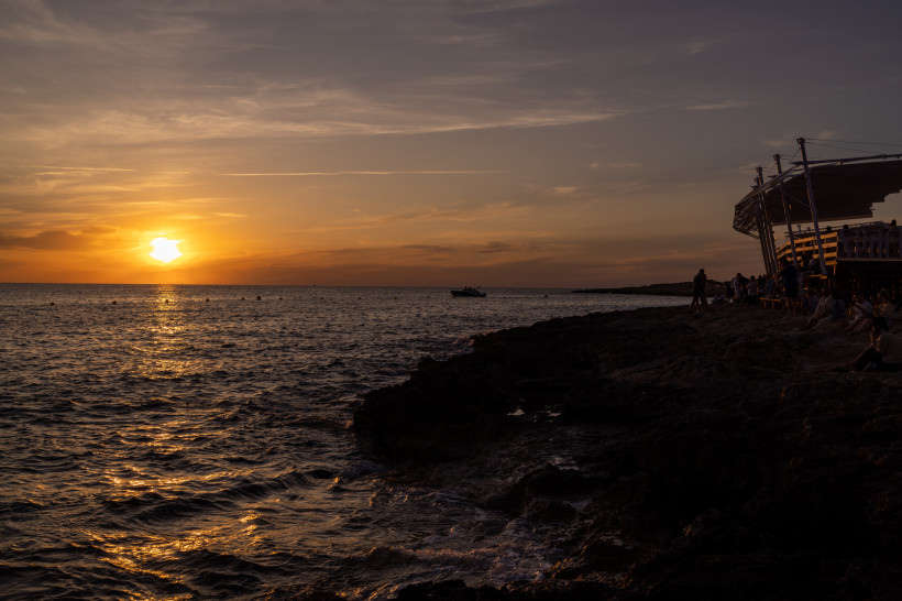 Sonnenuntergang über dem Meer bei Sant Antoni de Portmany, felsige Küste mit Menschen und Strandbar im Vordergrund