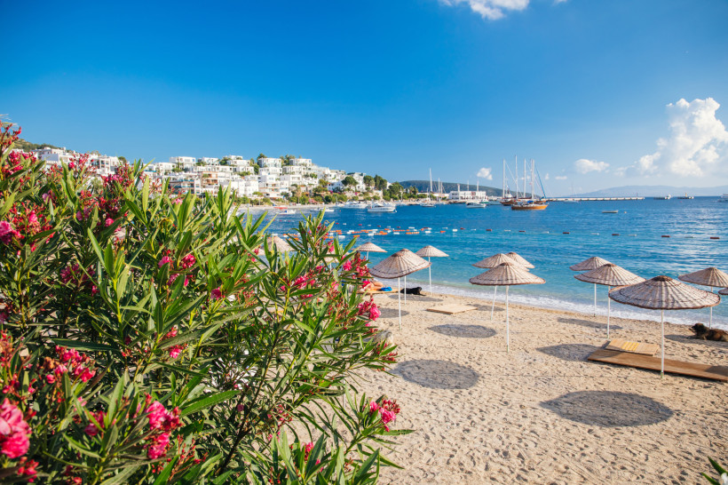 Sandstrand mit Sonnenschirmen, türkisblauem Meer und Yachten vor einer weißen Küstenstadt an der türkischen Ägäis