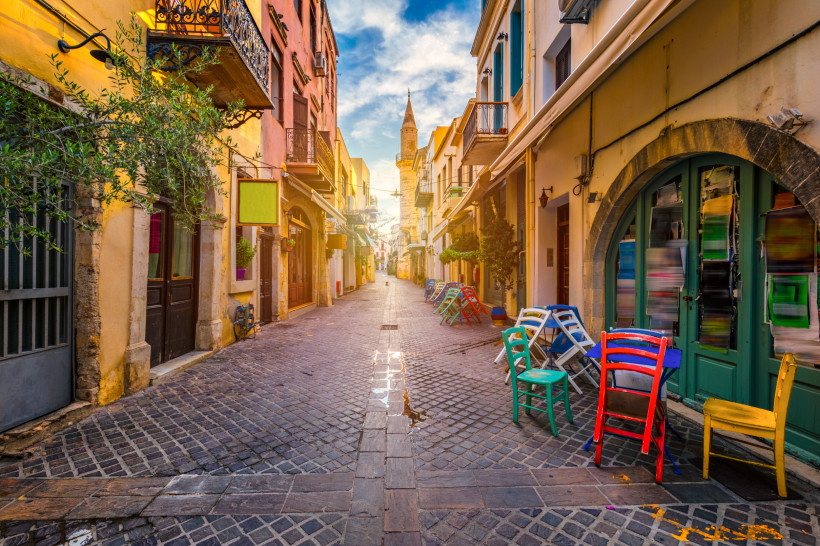 Gepflasterte Gasse in der Altstadt von Chania mit bunten Stühlen vor Gebäuden und Blick auf einen Turm