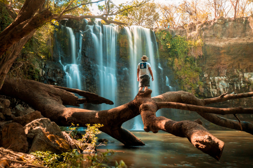 Costa Rica Ein Mann steht auf einem mächtigen umgestürzten Baumstamm, der über ein ruhiges Gewässer vor einem breiten Wasserfall ragt. Der Wasserfall stürzt in mehreren Kaskaden von einer Felskante herab, umgeben von dichtem Grün. Die Szene wirkt wild, abenteuerlich