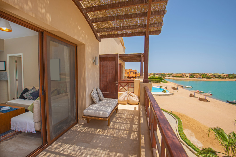Ägypten - El Gouna Blick von einem überdachten Balkon einer Ferienunterkunft direkt an einer Lagune in El Gouna, Ägypten. Auf dem Balkon befinden sich eine gepolsterte Sitzbank und Sitzsäcke. Rechts im Bild erstreckt sich ein ruhiger Sandstrand mit Blick auf türkisblaues Wa