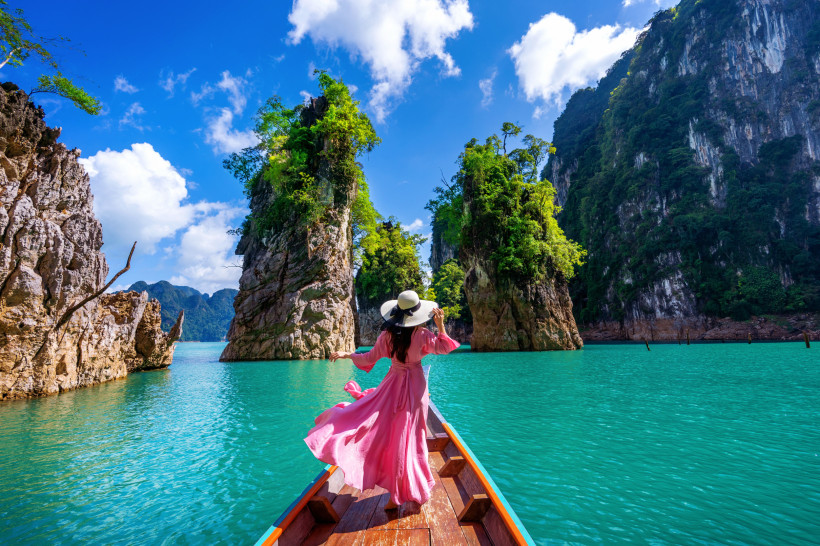 Frau in pinkem Kleid auf Langboot zwischen tropischen Felsen im türkisfarbenen Wasser unter blauem Himmel.
