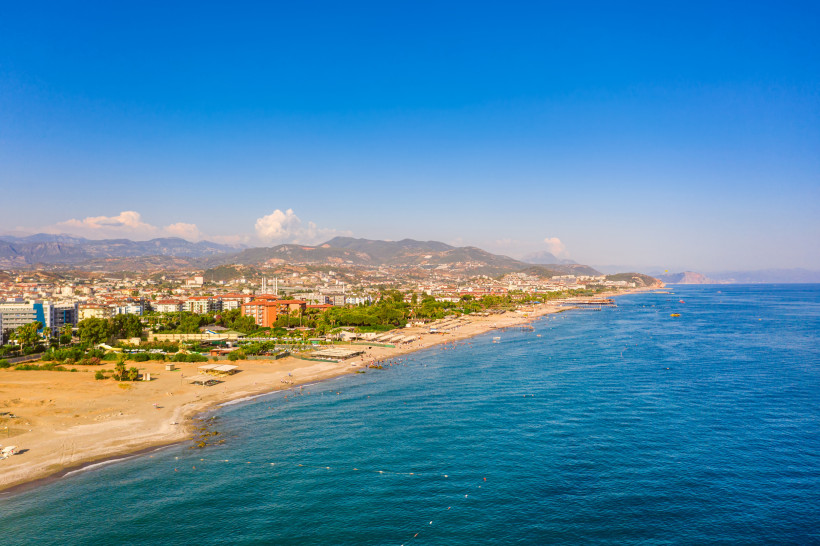 Das Bild zeigt einen langen, flach abfallenden Sandstrand in Konaklı. Entlang der Küste liegen Hotelanlagen und Wohnhäuser. Im Hintergrund erstrecken sich die Berge des Taurusgebirges und das tiefblaue Mittelmeer.