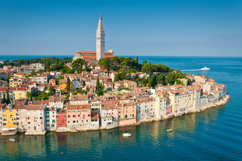 Blick auf die Altstadt von Rovinj in Kroatien. Bunte Häuser stehen dicht an dicht am Wasser, dahinter erhebt sich die barocke Kirche der Heiligen Euphemia mit ihrem hohen Glockenturm. Im Hintergrund das glitzernde Blau der Adria.