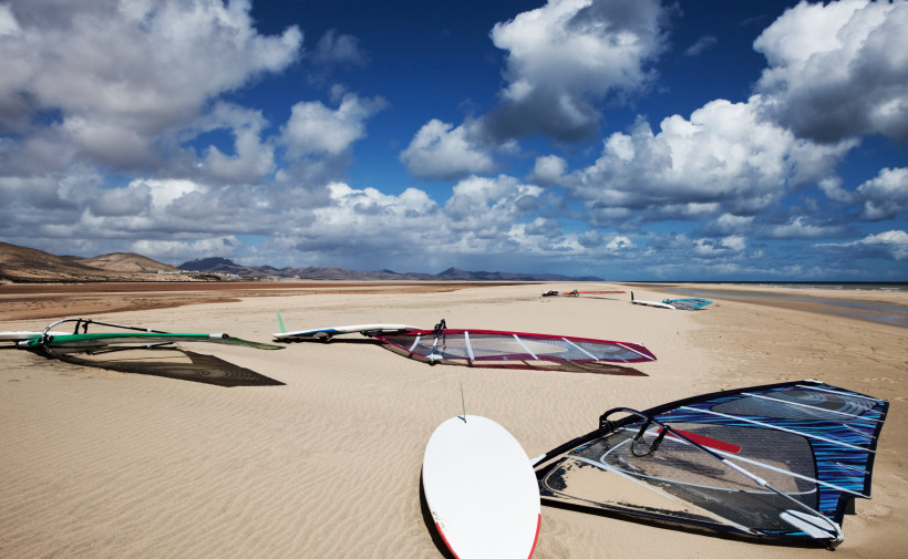 Fuerteventura Mehrere Windsurf-Boards mit Segeln liegen am sandigen Strand unter einem teils bewölkten Himmel. Im Hintergrund Berge und Weite der Küste