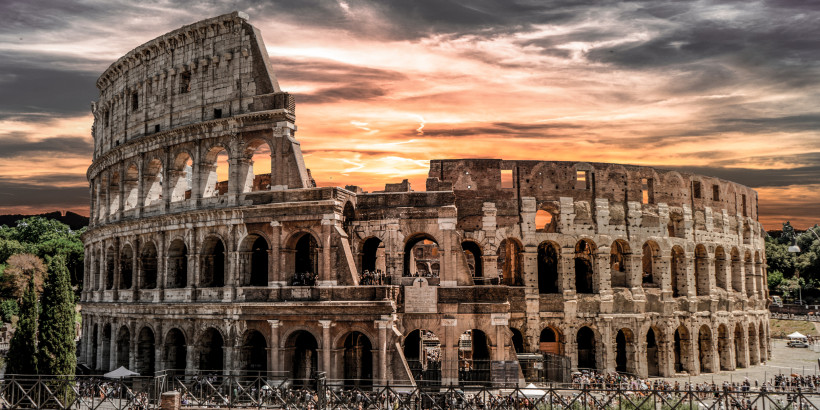Detailaufnahme des römischen Kolosseums in Rom bei dramatischem Sonnenuntergang. Die markanten Bögen der Arena leuchten in warmen Farben, während die untergehende Sonne den Himmel in Orange- und Rottönen färbt. Einige Besucher sind am unteren Rand zu sehe
