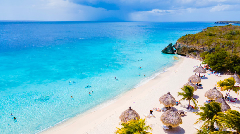 Sandstrand auf Curaçao mit türkisblauem Meer, Badegästen und Strohdach-Sonnenschirmen