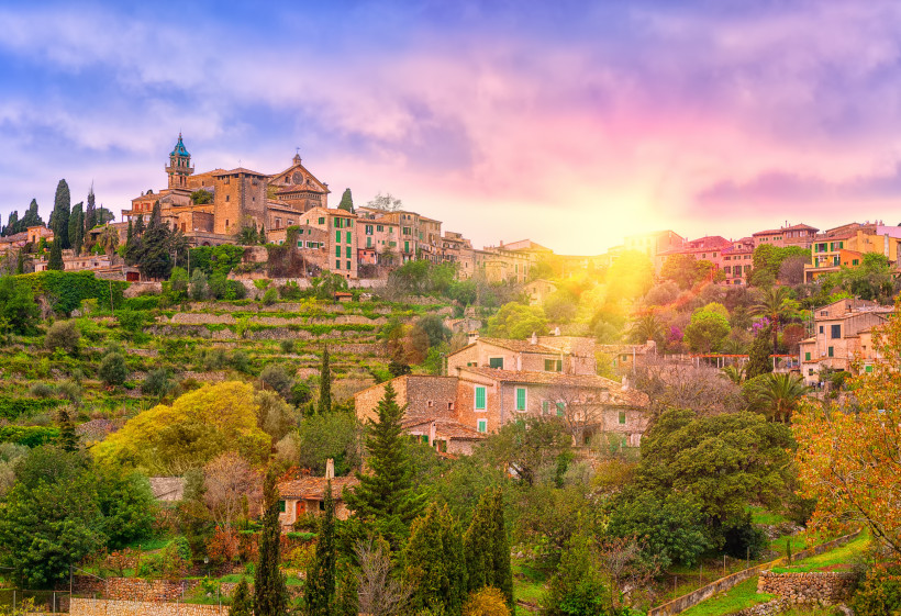 Mallorca Malerisches Bergdorf auf Mallorca bei Sonnenuntergang. Die historischen Steinhäuser mit grünen Fensterläden schmiegen sich an die terrassierten Hänge, umgeben von üppigem Grün, Zypressen und anderen Bäumen. Im oberen Teil des Bildes ist eine Kirche mit ei