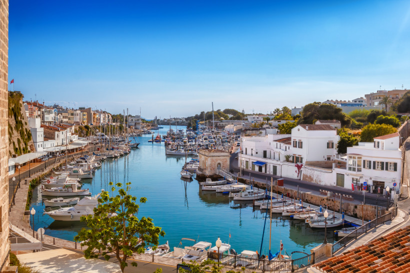 Blick auf den malerischen Hafen von Ciutadella auf Menorca, Spanien. Entlang des ruhigen, schmalen Wasserarms liegen zahlreiche Segelboote und Yachten vertäut. Auf beiden Seiten des Hafens reihen sich weiße und pastellfarbene Häuser, teils mit kleinen Caf