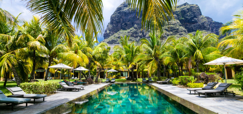 Mauritius Dieses Bild zeigt eine traumhafte Urlaubsidylle: Ein großes, elegantes Resort mit einem türkisfarbenen Infinity-Pool, der von hohen, sattgrünen Palmen eingerahmt wird. Schwarze Sonnenliegen mit weißen Sonnenschirmen laden zum Entspannen ein. Im Hintergrun
