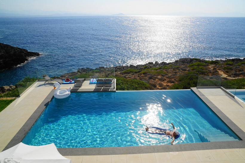 Kreta Ein Infinity-Pool mit Blick auf das offene Meer und eine felsige Küste. Eine Person treibt entspannt im Wasser, während eine aufblasbare Schwimmfigur in Form eines Schwans am Beckenrand schwimmt. Die Sonne spiegelt sich glitzernd auf der Wasseroberfläche,