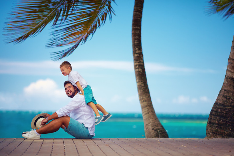 Lachender Vater in Sommerkleidung mit seinem Sohn auf den Schultern, am tropischen Strand mit Palmen und türkisblauem Meer