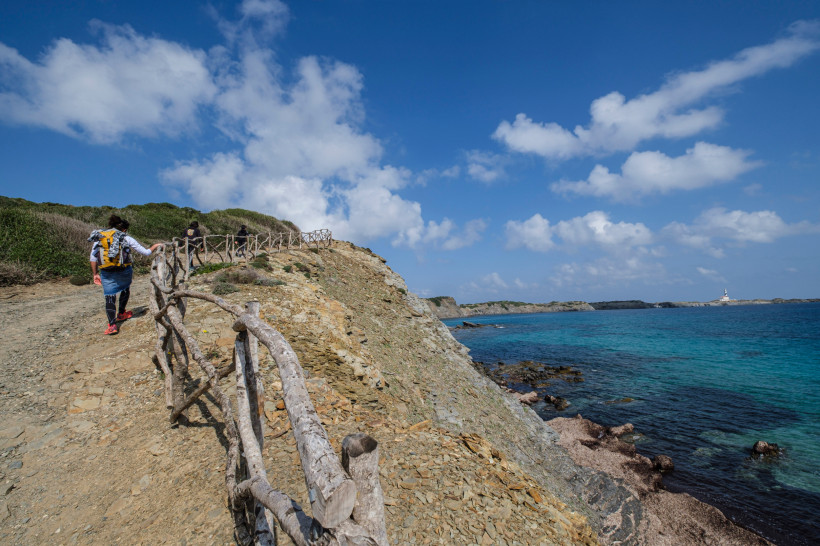 Mehrere Wanderer gehen einen schmalen Küstenpfad auf Menorca entlang, gesichert mit einem rustikalen Holzgeländer. Der Weg führt über eine felsige Anhöhe mit Blick auf das tiefblaue Meer. Am Horizont ist ein Leuchtturm zu sehen. Der Himmel ist mit weißen 