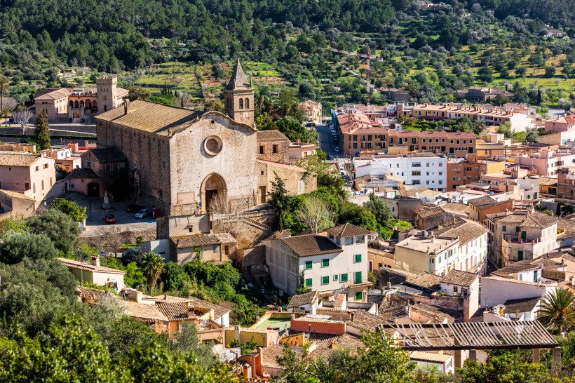 Mallorca Blick über ein malerisches Dorf mit dicht stehenden Häusern in warmen Farben und roten Dächern. Im Zentrum des Bildes eine große alte Kirche mit Rosettenfenster und einem Kirchturm. Im Hintergrund ein grünes Tal mit Feldern, Bäumen und vereinzelten Häuser