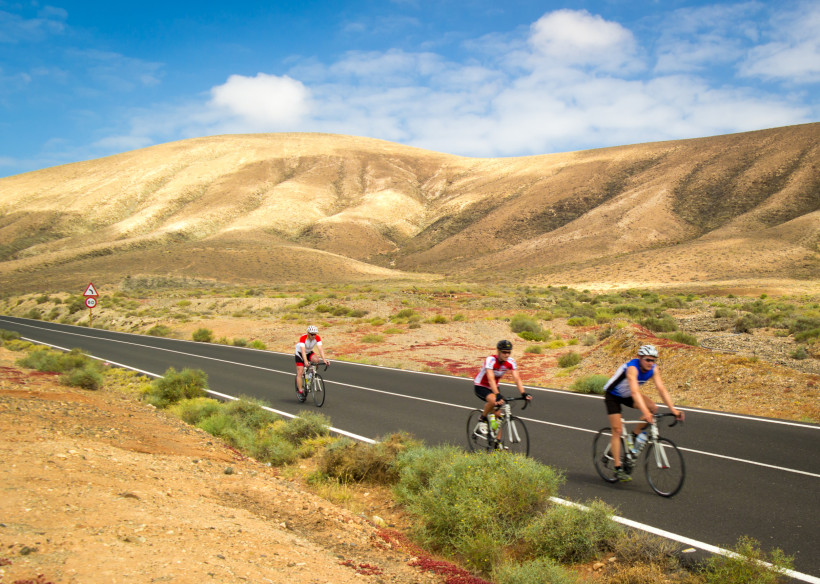 Drei Radfahrer fahren durch eine trockene, hügelige Vulkanlandschaft auf einer kurvenreichen Straße bei blauem Himmel