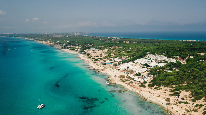 Formentera Luftaufnahme eines langen Strandes mit klarem, türkisfarbenem Wasser und einem Hotelkomplex im Grünen auf Formentera.