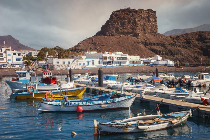 Hafen von Agaete – authentisches Fischerdorf auf Gran Canaria Boote schaukeln im Hafen von Agaete auf Gran Canaria vor der markanten Felsformation mit Kreuz auf dem Gipfel. Weiße Häuser, klares Wasser und maritimes Flair prägen die charmante Hafenatmosphäre.