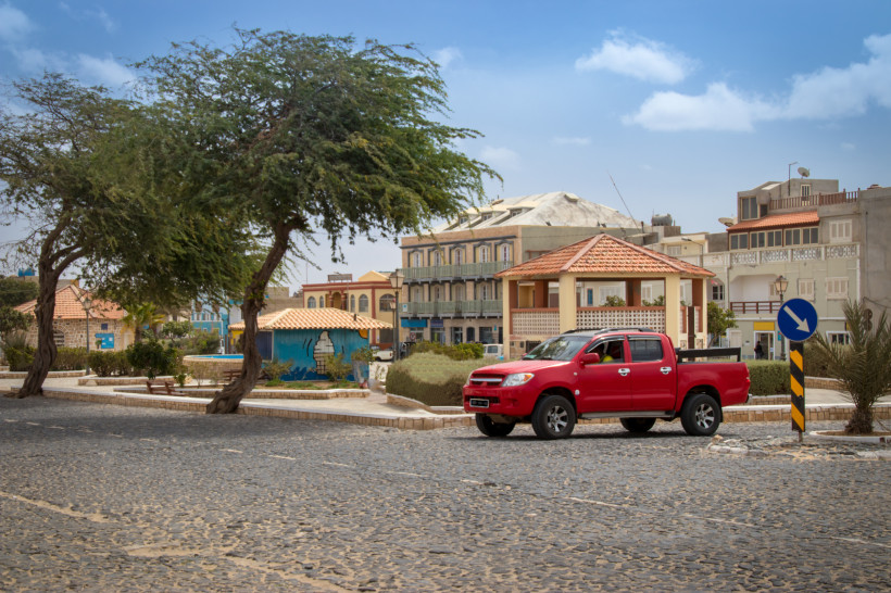 Straßenszene auf den Kapverden mit einem roten Pickup, der auf einer gepflasterten Straße an pastellfarbenen Häusern vorbeifährt. Im Hintergrund stehen moderne und traditionelle Gebäude, sowie ein bemaltes Wandbild mit maritimem Motiv.