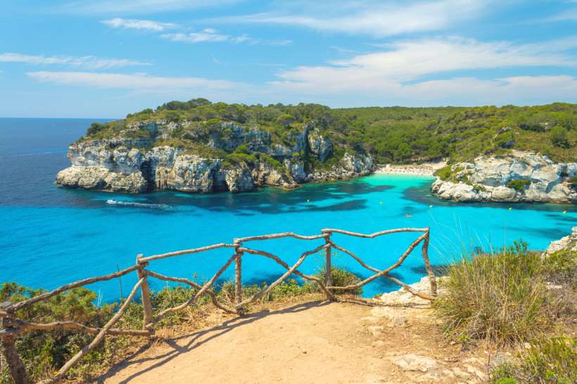 Atemberaubender Aussichtspunkt mit rustikalem Holzgeländer über einer türkisblauen Bucht auf Menorca. Im Hintergrund liegen helle Kalkfelsen, grüne Pinienwälder und ein kleiner Sandstrand mit Badenden. Das Wasser leuchtet in kräftigem Blau, ein Boot hinte