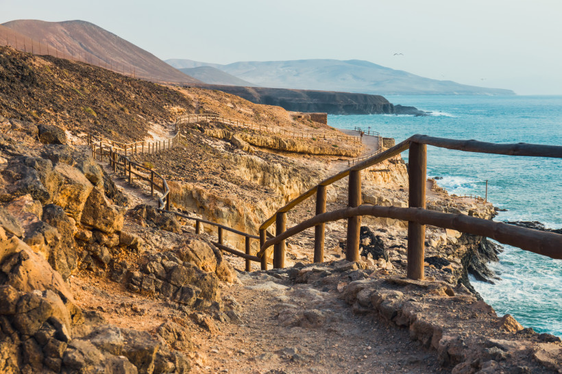 Wanderweg entlang der felsigen Küste von Fuerteventura mit Holzgeländer und Blick auf den Atlantik.
