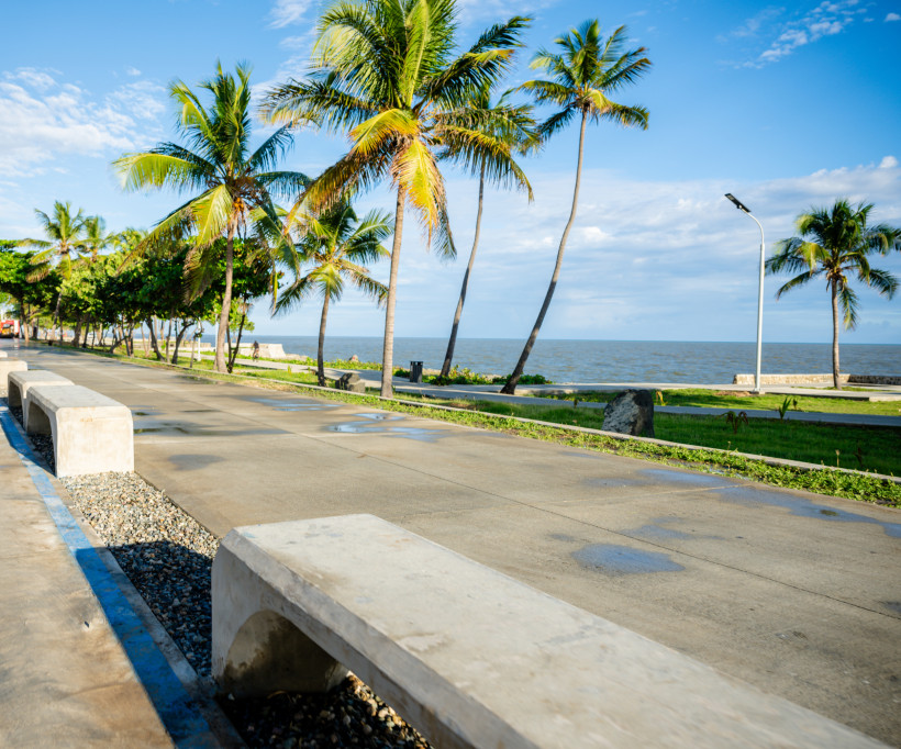 Ruhige Promenade mit Betonbänken und Palmen direkt am Meer unter einem klaren Himmel, mit vereinzelten Wasserpfützen auf dem Weg