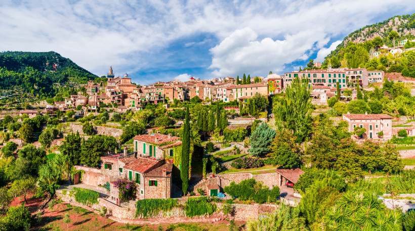 Mallorca - Valldemossa Ein malerischer Blick auf ein mediterranes Dorf auf Mallorca, das sich an einen Hang schmiegt. Die Häuser bestehen aus Naturstein mit grünen Fensterläden und sind von Zypressen, Pinien und anderen mediterranen Pflanzen umgeben. Der Hintergrund zeigt einen