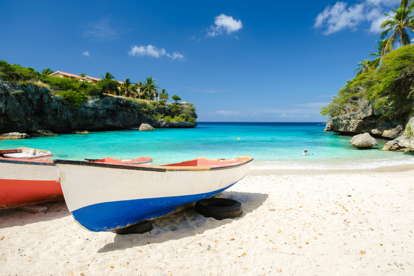 Curaçao Zwei kleine Fischerboote stehen auf dem hellen Sandstrand einer türkisblauen Bucht. Die Boote sind weiß lackiert mit roten und blauen Akzenten. Im Hintergrund ragen grüne Felsen ins Meer, darüber Palmen und gelb gestrichene Häuser mit roten Dächern. Das r