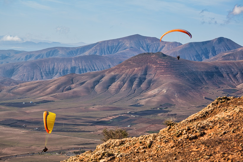 Zwei Gleitschirmflieger schweben über eine vulkanische Berglandschaft mit klarer Sicht und ruhigem Himmel