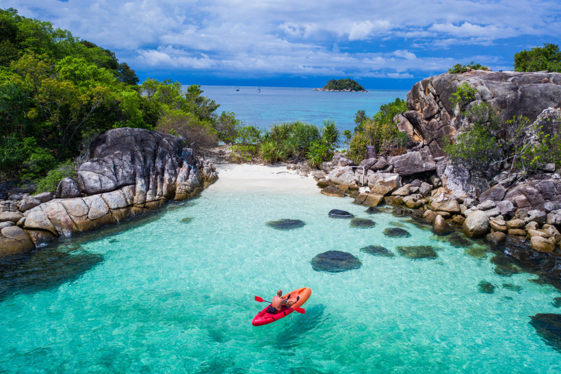 Thailand Ein Mann paddelt mit einem roten Kajak durch eine türkisfarbene, tropische Bucht. Umgeben von Felsen, grüner Vegetation und glasklarem Wasser führt der Weg auf eine kleine, abgelegene Sandbucht zu. Im Hintergrund sind Segelboote und eine kleine Insel zu s