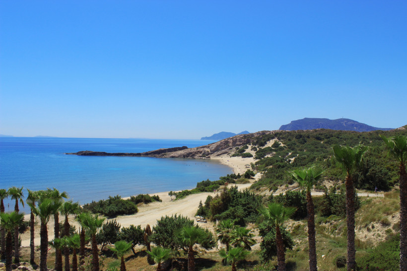 Kos Das Bild zeigt eine ruhige, geschwungene Bucht mit einem breiten Sandstrand. Im Vordergrund stehen zahlreiche Palmen und grüne Sträucher. Der Sandstrand erstreckt sich entlang des klaren, türkisblauen Wassers. Auf dem Strand sind vereinzelt Menschen zu er