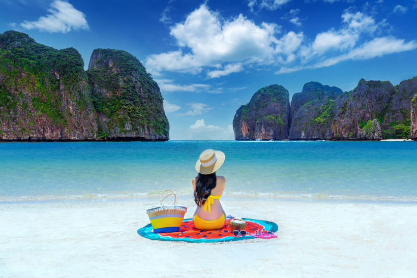 Frau sitzt am weißen Sandstrand der Maya Bay auf den Phi Phi Inseln bei Phuket, Thailand, mit Blick auf türkisfarbenes Meer und Kalksteinfelsen.