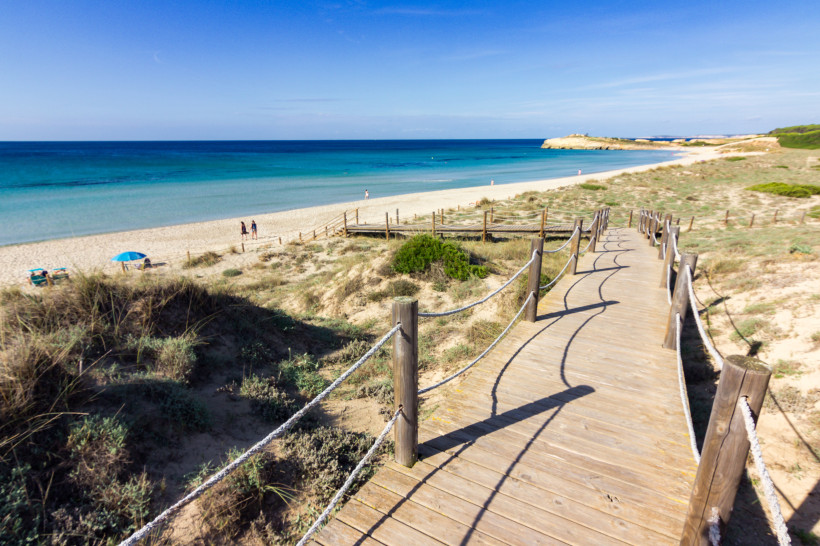 Menorca - Son Bou Ein hölzerner Steg mit Seilen führt über Dünen zum breiten Sandstrand von Son Bou auf Menorca. Im Hintergrund erstreckt sich das türkisfarbene Mittelmeer.