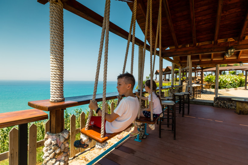  Pause mit Aussicht auf Korfu: Zwei Kinder genießen den Blick aufs Meer von Schaukeln aus, die an einer gemütlichen Holzterrasse befestigt sind. Das Café ist liebevoll mit Stein und Holz gestaltet. Der Ausblick reicht über das türkisfarbene Wasser bis zum
