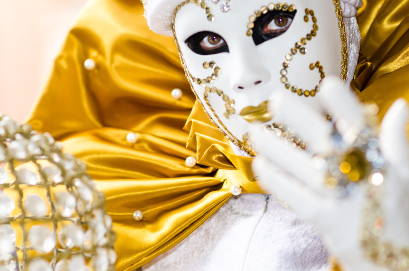 Nahaufnahme einer weiß-gold verzierten Maske mit glitzernden Steinchen. Die Maske wird von einer Person getragen, die in ein prächtiges Kostüm aus glänzendem goldfarbenem Stoff mit Perlen gehüllt ist. Der Blick ist geheimnisvoll und ausdrucksstark.