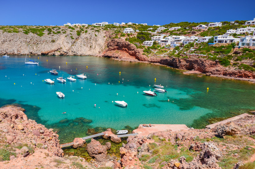 Kleine malerische Bucht auf Menorca mit türkisfarbenem Wasser und zahlreichen weißen Booten. Auf dem Hang entlang der Bucht stehen viele weiße Ferienhäuser mit Blick aufs Meer. Die Felsenküste ist rötlich gefärbt und kontrastiert mit dem klaren Wasser. Im