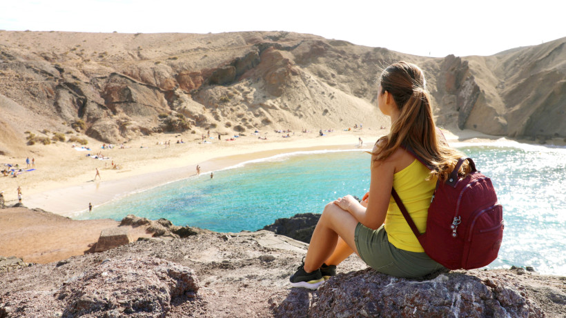 Playa de Papagayo Lanzarote – Traumstrand für Alleinreisende Alleinreisende Frau sitzt oberhalb des Playa de Papagayo und blickt auf den goldenen Strand und das türkisfarbene Meer von Lanzarote – ideal für entspannte Erkundungen.