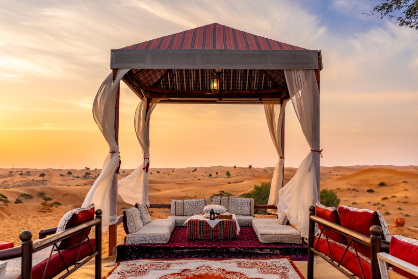 Arabisches Wüstencamp bei Sonnenuntergang mit einem offenen Zeltpavillon in traditionellem Design. Im Inneren liegen orientalische Teppiche und gemütliche Sitzpolster rund um einen niedrigen Tisch. Leichte Vorhänge wehen im Wind, im Hintergrund erstreckt 