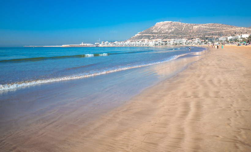 Weiter Sandstrand von Agadir mit sanften Wellen und türkisblauem Wasser. Im Hintergrund weiße Gebäude der Stadt und ein Hügel mit arabischer Schrift, unter klarem, wolkenlosem Himmel.