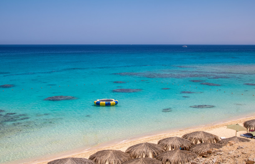 Ägypten - Hurghada Ein türkisfarbenes, klares Meer mit flachen Korallenbänken, in dem eine bunte Wassertrampolin-Insel schwimmt. Im Vordergrund sind strohgedeckte Sonnenschirme am Sandstrand zu sehen.