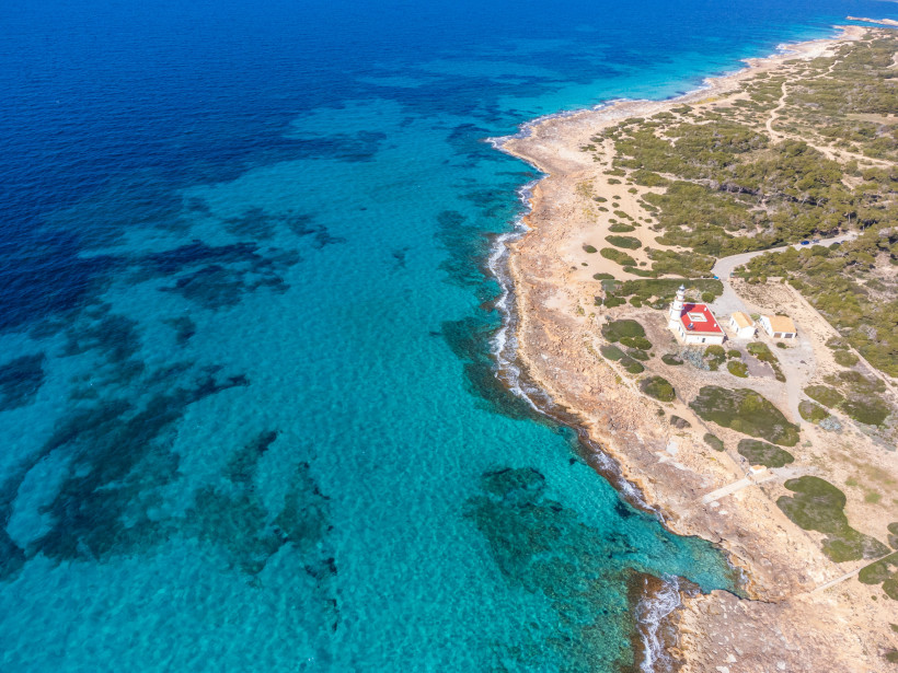 Mallorca - Cap de Ses Salines Eine felsige Küste mit einem markanten Leuchtturm mit rot-weißem Dach, umgeben von flachen Sträuchern und Sandpfaden. Das Meer ist türkisblau und trifft auf die zerklüfteten Felsen der Küste. Der Leuchtturm steht auf einem leicht erhöhten Plateau und dien