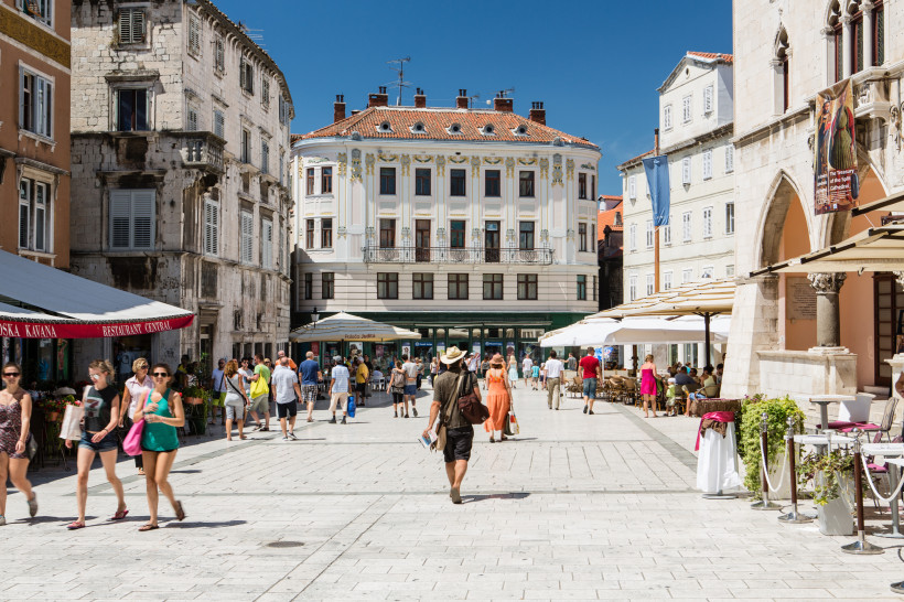 elebter Platz in der Altstadt von Split mit historischen Gebäuden, Straßencafés und vielen flanierenden Menschen bei Sonnenschein. Im Hintergrund ein prachtvolles Gebäude mit verzierten Fensterrahmen.