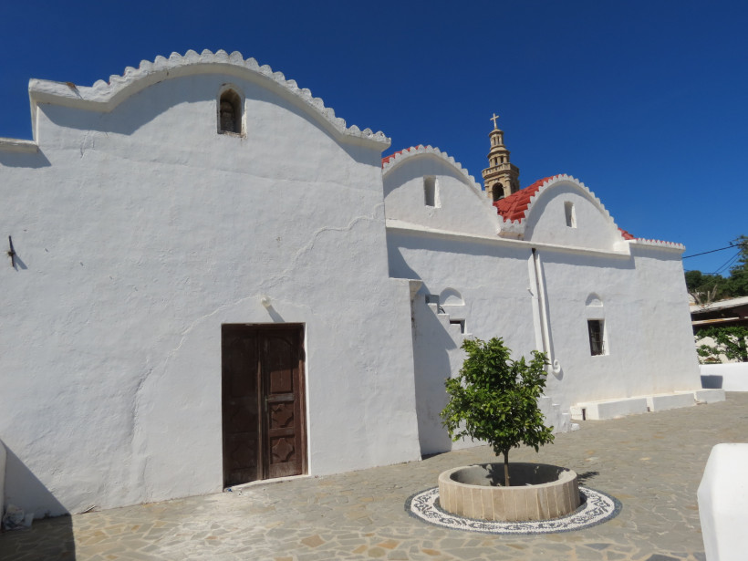 Weiße griechische Kirche mit geschwungenem Dach, rotem Ziegeldach und Glockenturm unter tiefblauem Himmel auf Rhodos