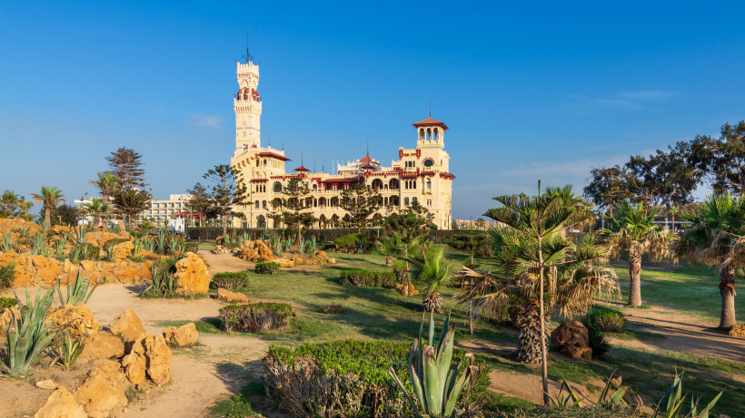 Das Bild zeigt den prächtigen Montaza Palast in Alexandria, Ägypten. Das Schloss im neoklassizistisch-orientalischen Stil mit hohen Türmen, roten Dächern und verzierten Fassaden steht im Hintergrund. Davor erstreckt sich eine gepflegte Parklandschaft mit 