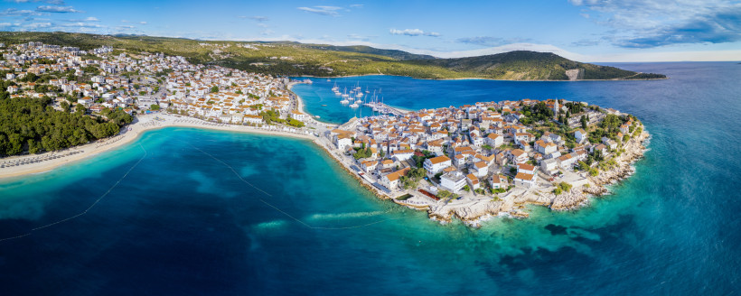 Kroatien Panoramablick auf eine Küstenstadt mit dicht gedrängten Häusern mit roten Dächern auf einer Halbinsel. Rechts und links davon strahlen türkisblaues Meer und mehrere Yachten im Hafen. Im Hintergrund bewaldete Hügel.