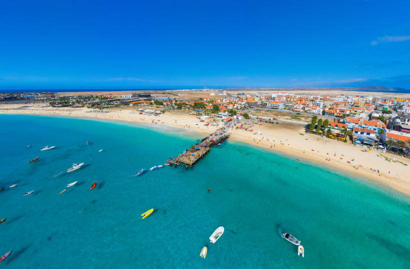 Luftaufnahme eines weiten Sandstrandes mit kristallklarem, türkisblauem Wasser auf der Kapverden-Insel Sal. Am Strand liegen Sonnenanbeter, im Wasser sind zahlreiche kleine Boote. Ein langer Holzsteg ragt ins Meer, am Ufer stehen bunte Gebäude, Hotels und