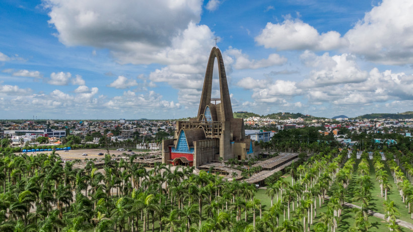 Luftaufnahme der monumentalen Basilika Unserer Lieben Frau von Altagracia in Higüey, umgeben von hohen Palmen, mit Blick auf die Stadt unter einem bewölkten Himmel