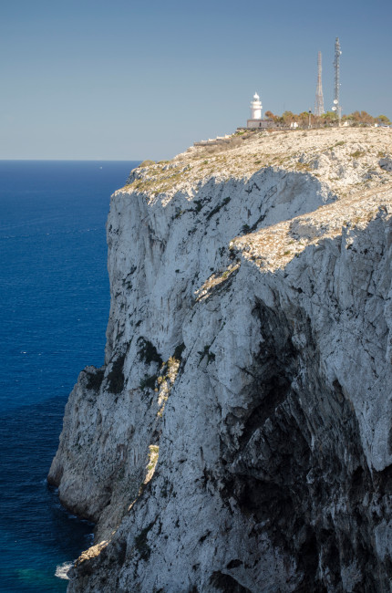 Mallorca Ein weißer Leuchtturm steht auf einer imposanten, steil abfallenden Kalkstein-Klippe über dem tiefblauen Mittelmeer. Daneben befinden sich Funkmasten und einige Bäume. Die Klippe wirkt gewaltig und zerklüftet, während das Meer ruhig und grenzenlos in den