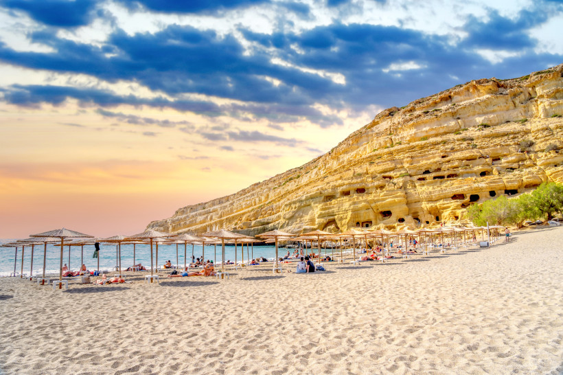 Kreta Ein breiter Sandstrand mit Liegen und Strohdächern liegt vor einer eindrucksvollen Felswand mit Höhlenöffnungen. ( Matala ) Menschen entspannen unter den Schirmen, während am Himmel ein farbenfroher Sonnenuntergang mit dunklen Wolken zu sehen ist.