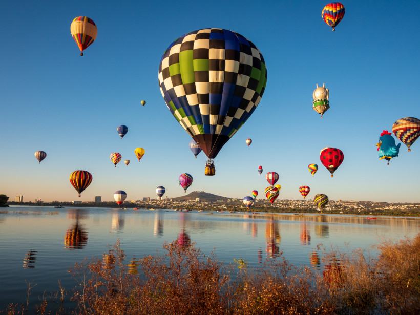 Internationales Heißluftballon-Festival in Leon Mehrere bunte Heißluftballons steigen am frühen Morgen über einem See mit ruhigem Wasser auf, während die Stadt und Hügel im Hintergrund sichtbar sind